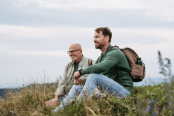 Adult son and older father hiking togeter in nature, resting on meadow. Spending time together.